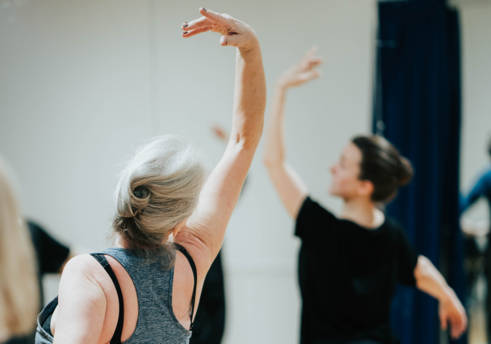A person stands with their back to the camera, their arm is stretched elegantly above their head in a ballet pose. They are looking towards a ballet instructor, who is out of focus in the background but is doing the same pose.