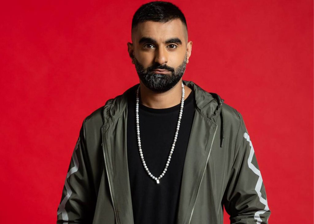 Tez Ilyas looking towards camera wearing green jacket on red background