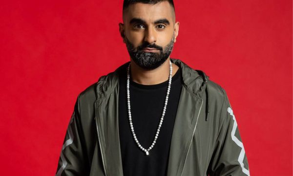 Tez Ilyas looking towards camera wearing green jacket on red background