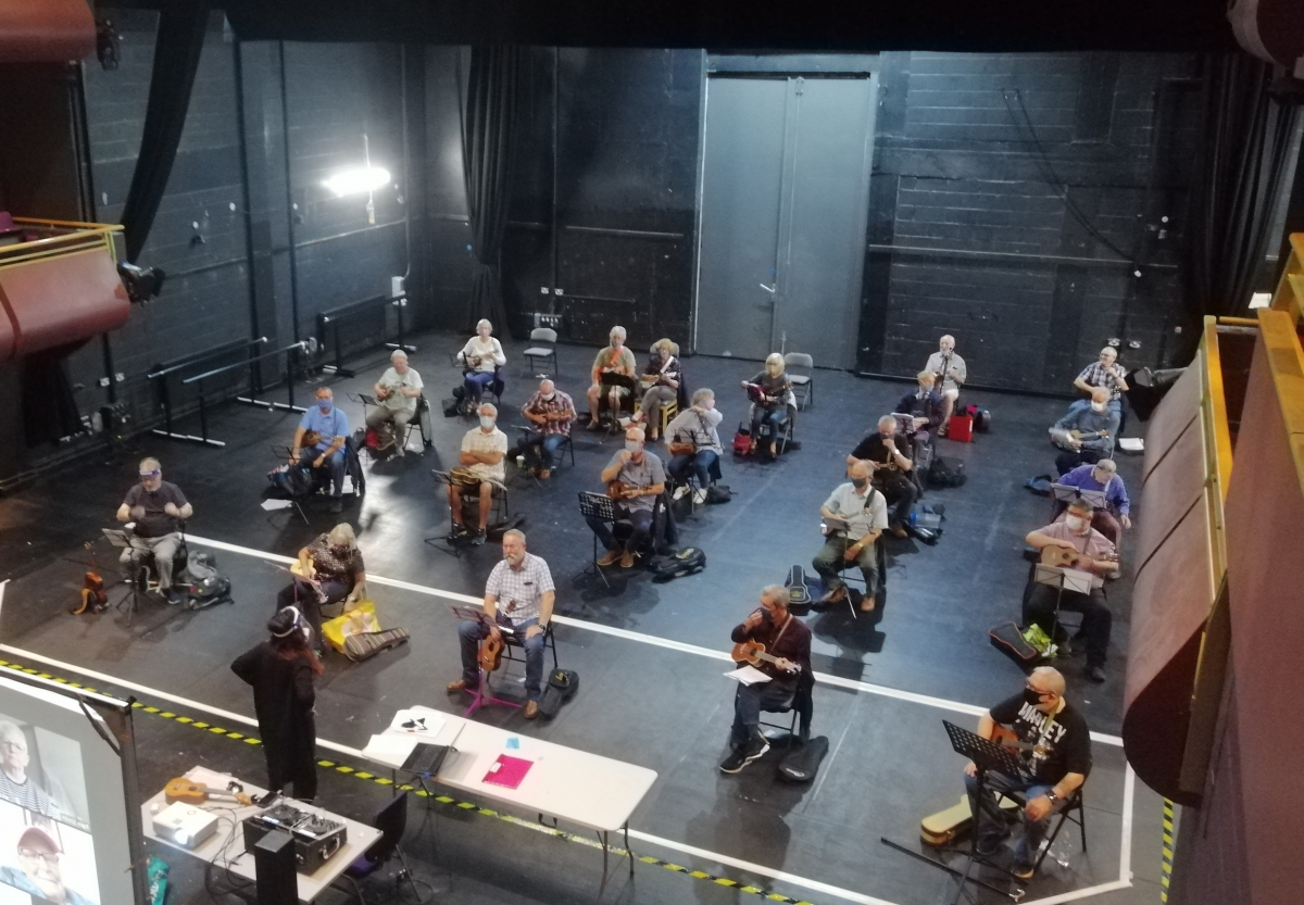 Image of a group of people sat socially distanced on a theatre stage holding ukuleles, at the front a woman stands demonstrating something to the group, there are also a number of people projected on a screen behind her, who have joined by Zoom.