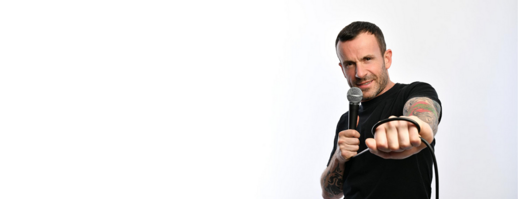 Comedian Gary Meikle (short dark hair and tattooed arms in a black t-shirt) holds a microphone in a cheeky pose