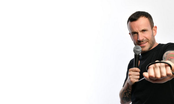 Comedian Gary Meikle (short dark hair and tattooed arms in a black t-shirt) holds a microphone in a cheeky pose