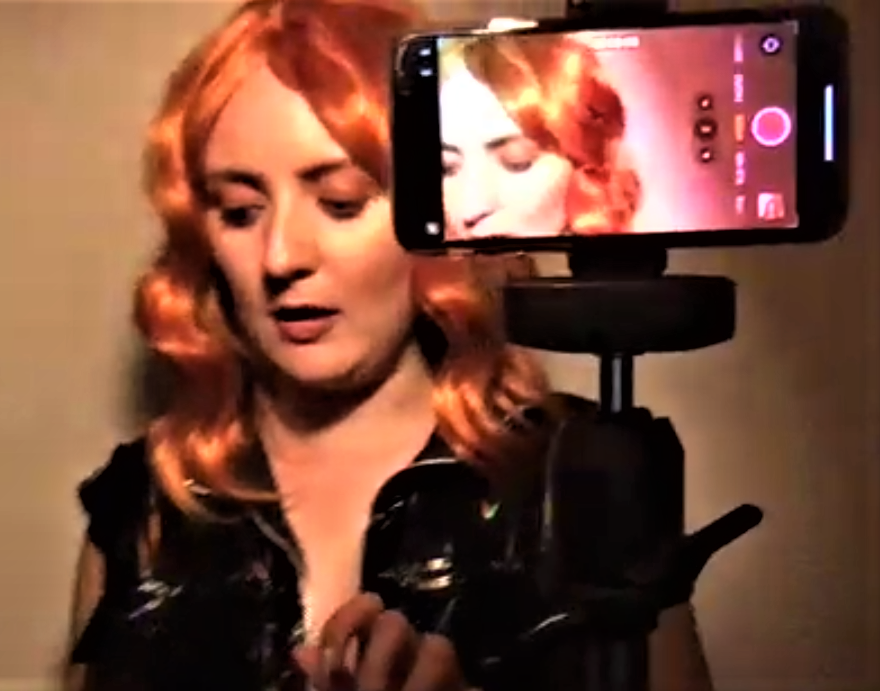 Philippa Cole is playing the character of Mim in a filmed scene for Siege. She is wearing an orange costume wig and a PVC dress and there is a second image of her off to the side viewed through a phone camera on a tripod
