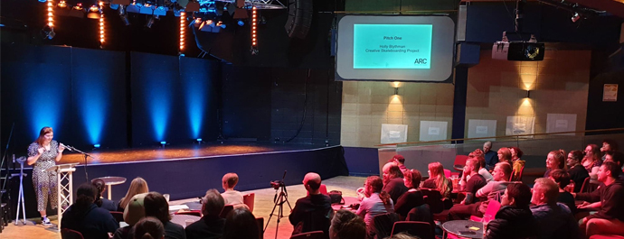 A person stands behind a lectern, pitching their project idea to an audience in The Point at ARC.