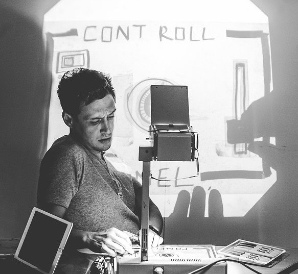 Black and white image of Scott Turnbull sitting behind an overhead projector. An illustration he;s creating is projected onto a screen behind him.