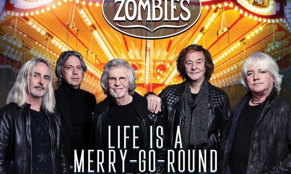 The rock band The Zombies stand in front of a brightly lit merry go round. They all look into the camera. Text in front of them reads "Life Is A Merry Go Round Tour."