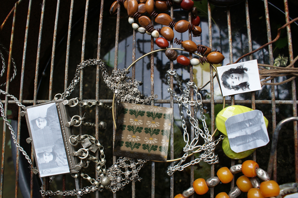 A selection of beads, other jewellery and photographs (of people) lie on a metal grille over water.