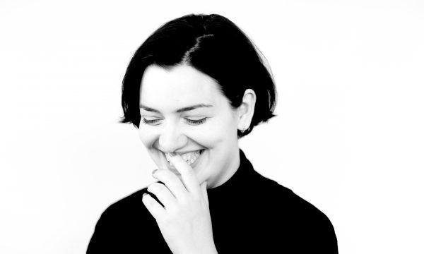 Black and white headshot of Lauren Vevers. Lauren is looking downwards and smiling, and has one hand to her mouth.