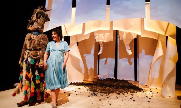A man and woman stand on stage in front of a set created from paper and coal. The woman is wearing a sky blue dress and the man is dressed in costume.