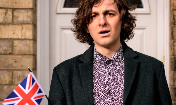 A man with a slightly confused look on his face, waving a small Union Jack, stands in front of a white front door