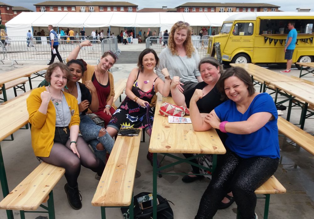 A group of the Tees Women Poets seated at tables and benches and posing for their photo each with one arm in a bodybuilding style pose, to show off their power.