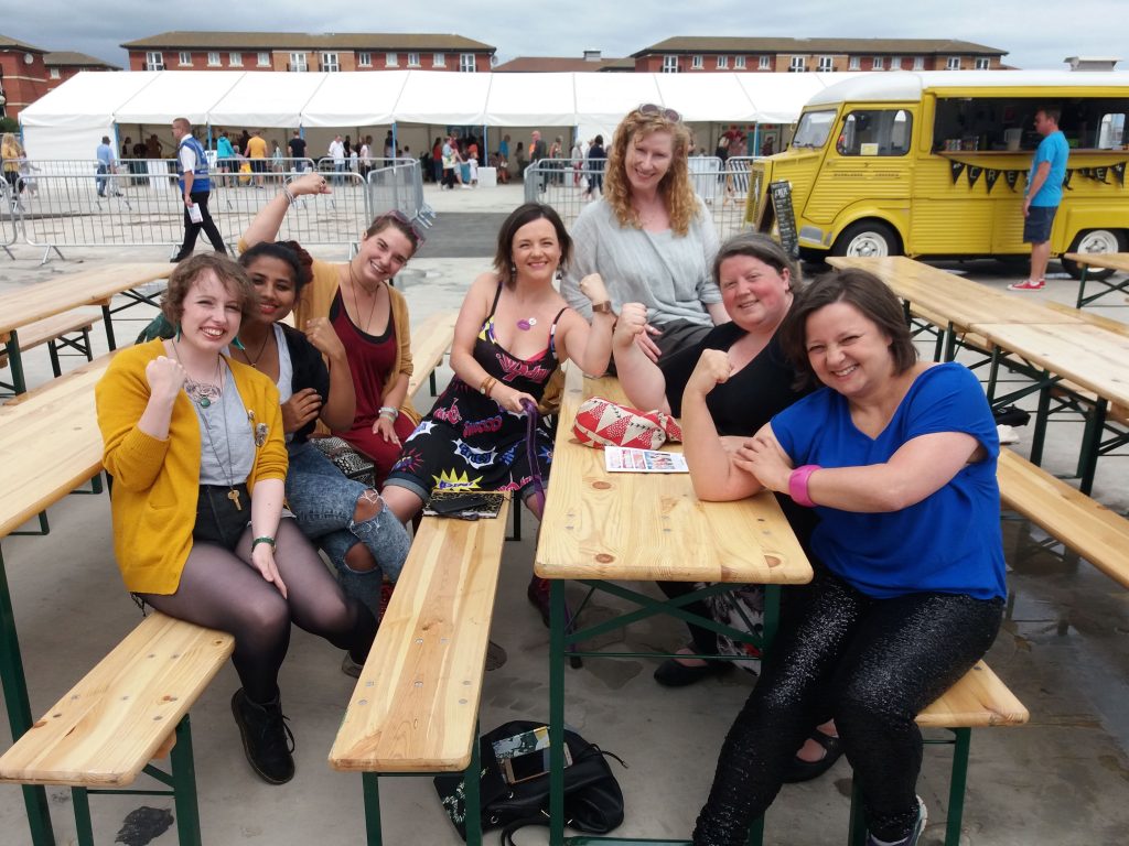 A group of the Tees Women Poets seated at tables and benches and posing for their photo each with one arm in a bodybuilding style pose, to show off their power.
