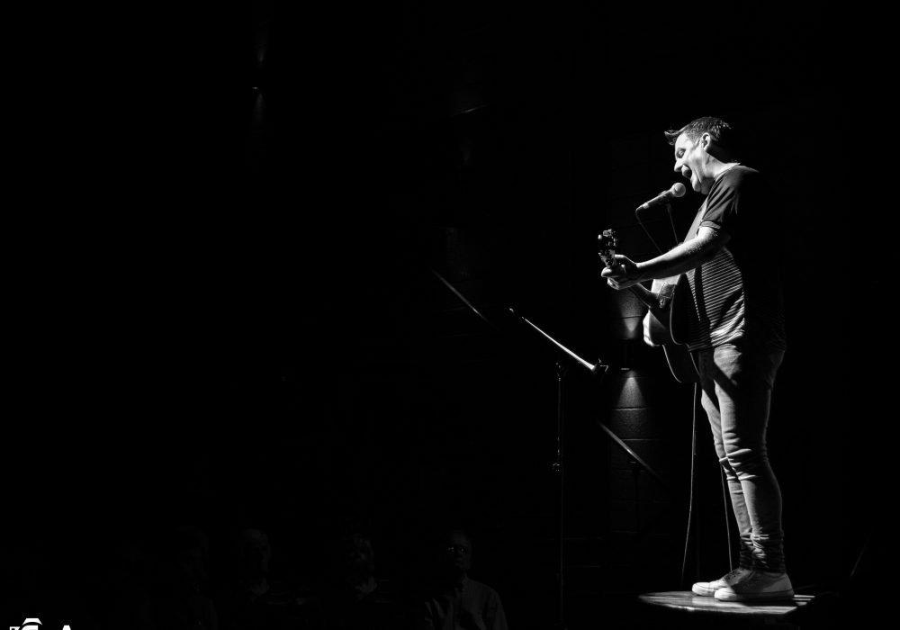 Black and White photo of Mike McGrother standing on a table in a dark room. He has a guitar and is singing into a microphone. by EnA Photography