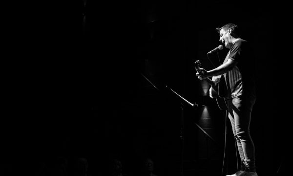 Black and White photo of Mike McGrother standing on a table in a dark room. He has a guitar and is singing into a microphone. by EnA Photography