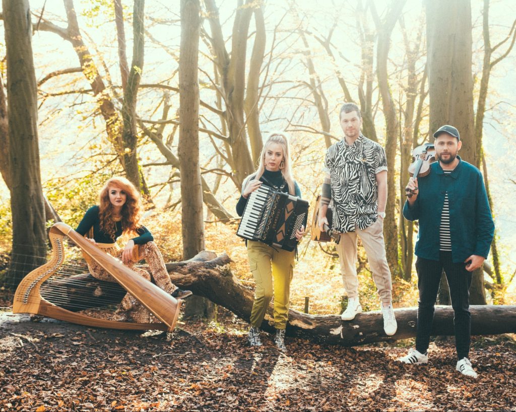 Folk band Calan stand in a golden tinged forest. On the left is a red-haired harpist sitting on top of a fallen tree trunk, an accordian player holds her instrument, and a guitarist and violinist are both standing beside the same fallen tree.