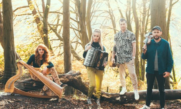 Folk band Calan stand in a golden tinged forest. On the left is a red-haired harpist sitting on top of a fallen tree trunk, an accordian player holds her instrument, and a guitarist and violinist are both standing beside the same fallen tree.