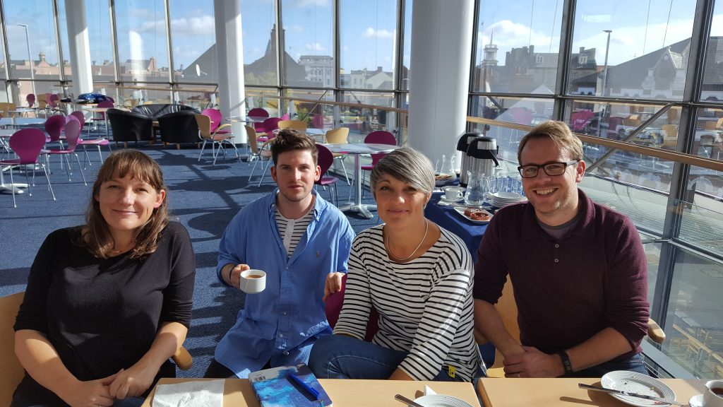 Four members of Fidget Theatre sitting at a table in ARC's second floor gallery space on a sunny day, with blue skies visible through the large curved glass windows behind them