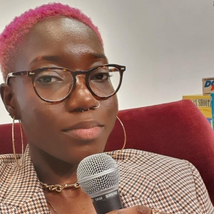 Journals of Dami - a black woman with short pink hair and glasses, holding a microphone