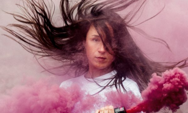Luca stands facing the camera surrounded by pink smoke, and her long dark hair flying around her head