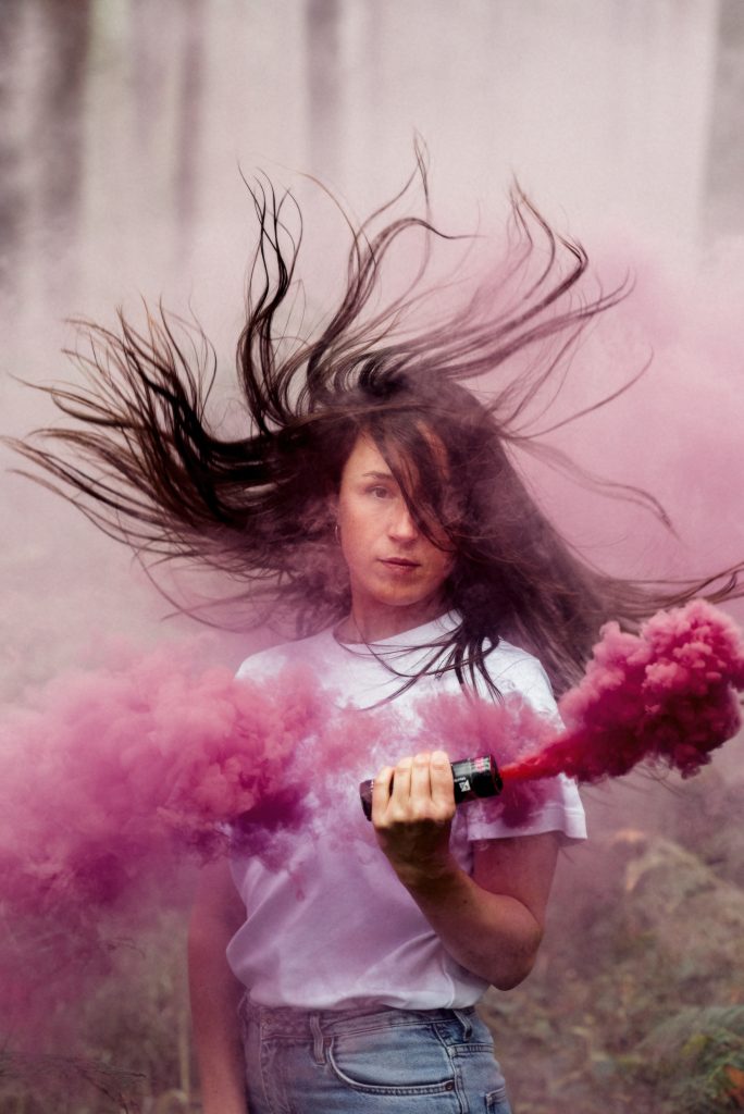 Luca stands facing the camera surrounded by pink smoke, and her long dark hair flying around her head