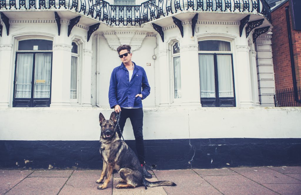 Poet Luke Wright, dressed in dark blue jeans, a white t-shirt, a blue shirt and sunglasses, stands in front of a white building holding a German Shepherd dog on a lead