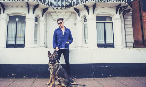 Poet Luke Wright, dressed in dark blue jeans, a white t-shirt, a blue shirt and sunglasses, stands in front of a white building holding a German Shepherd dog on a lead