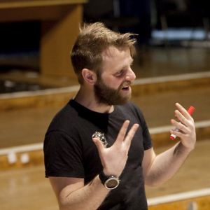 Headshot of artist Barra Collins speaking while gesturing with his hands, and holding a pen in his hand