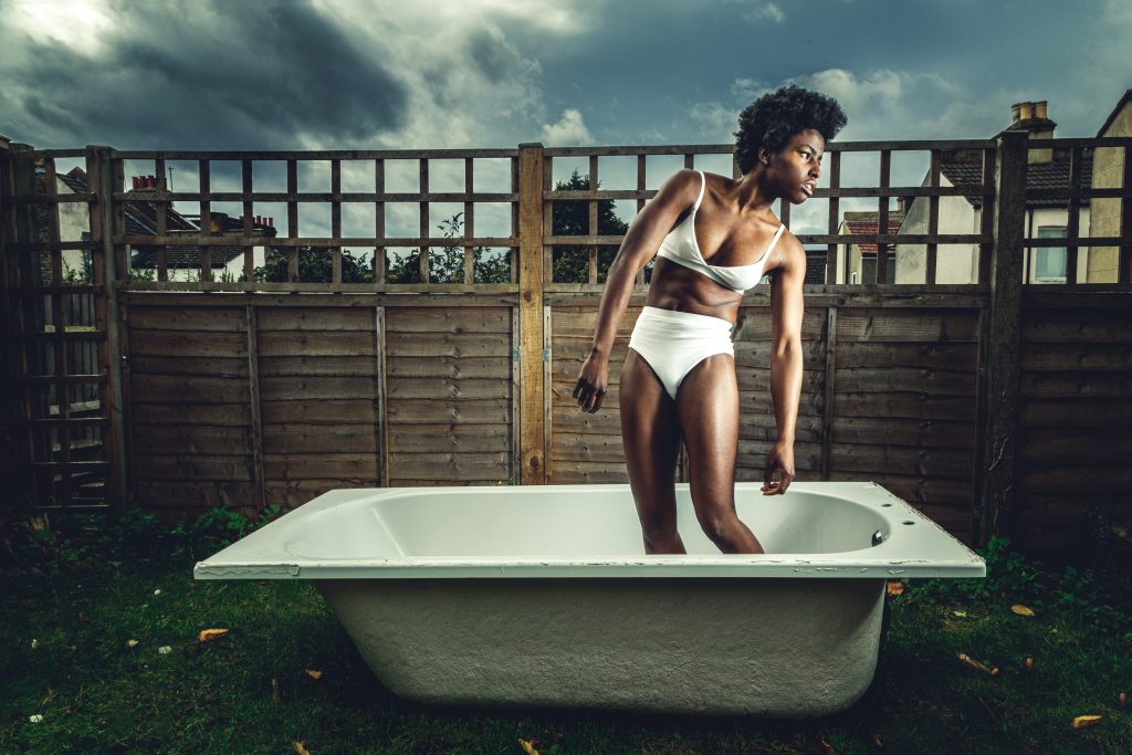 Project image of Farrell Cox, a black woman wearing a white bikini, standing in a bath in a garden.