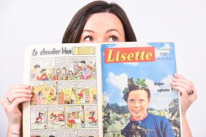 Lisette, a white woman with freckles and brown hair, peers over the top of a colourful French comic that is called 'Lisette'.