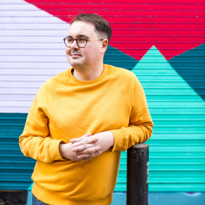 Image of artist Tommy, wearing round glasses and a bright yellow jumper, in front of brightly, multi-coloured painted shutters