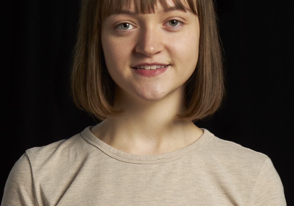 Headshot of Niamh Nelson. She is in her early 20s and has mid-brown straight haior that falls just under chin length. She is wearing a short-sleeve, beige coloured top.
