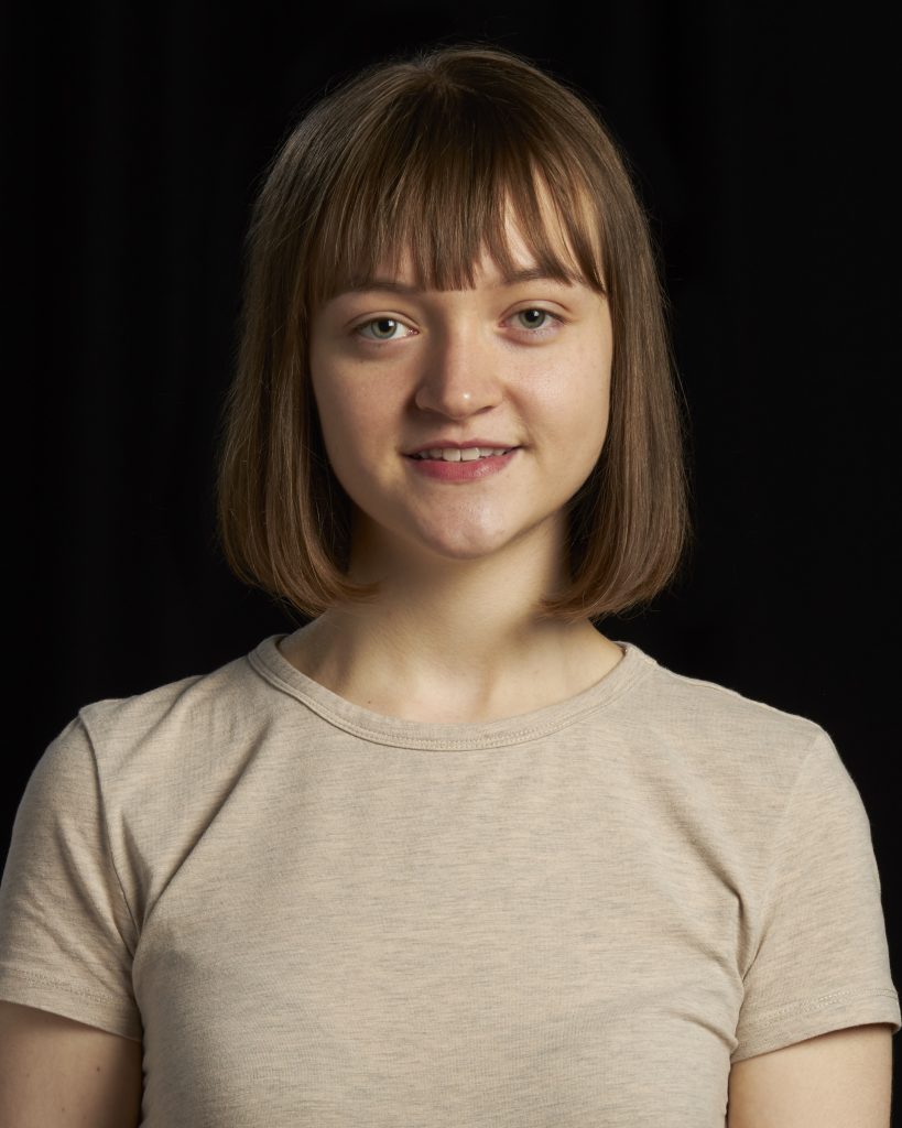 Headshot of Niamh Nelson. She is in her early 20s and has mid-brown straight haior that falls just under chin length. She is wearing a short-sleeve, beige coloured top.