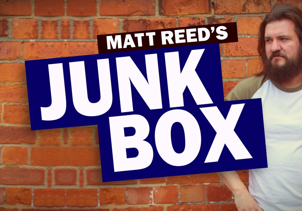 Comedian Matt Reed (shoulder length straight dark hair, beard, wearing a white top with green sleeves) is standing outside against a red brick wall. Text overlaid on the image reads 'MATT REED'S JUNK BOX'