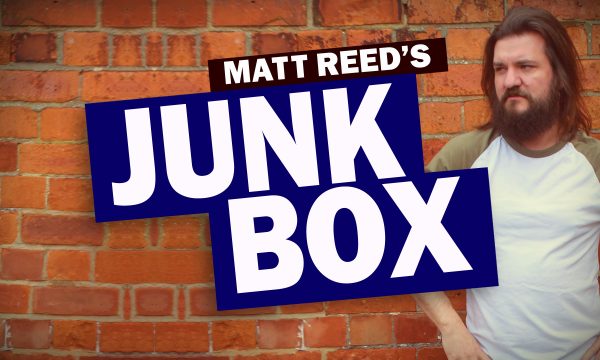 Comedian Matt Reed (shoulder length straight dark hair, beard, wearing a white top with green sleeves) is standing outside against a red brick wall. Text overlaid on the image reads 'MATT REED'S JUNK BOX'