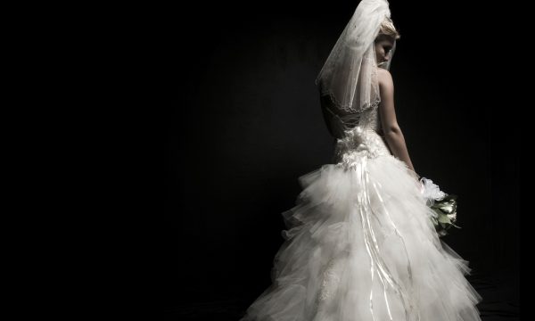 A woman in a white wedding dress and veil, seen from behind, on a black background