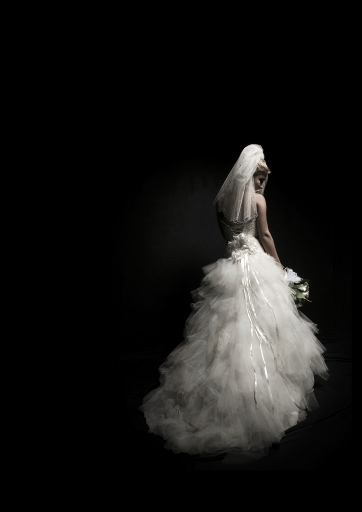 A woman in a white wedding dress and veil, seen from behind, on a black background