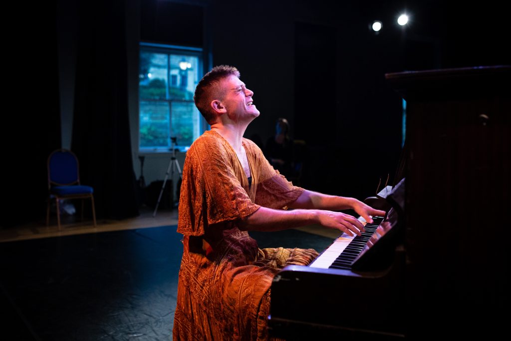 Mike Auger from Displace Yourself Theatre sits playing a piano. He is wearing a loose, orange, Indian inspired outfir.