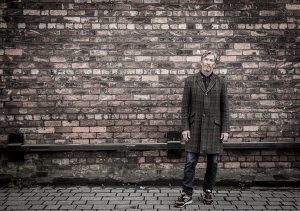 Mike Garry, wearing an overcoat, stands against a plain brick wall