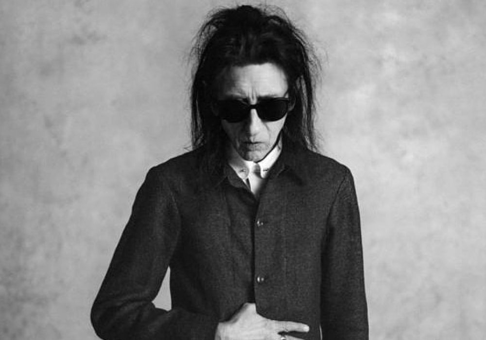 Black and white image of John Cooper Clarke. He has long dark hair, and is wearing sunglasses and a dark jacket buttoned almost to the top. He is looking down towards the floor.