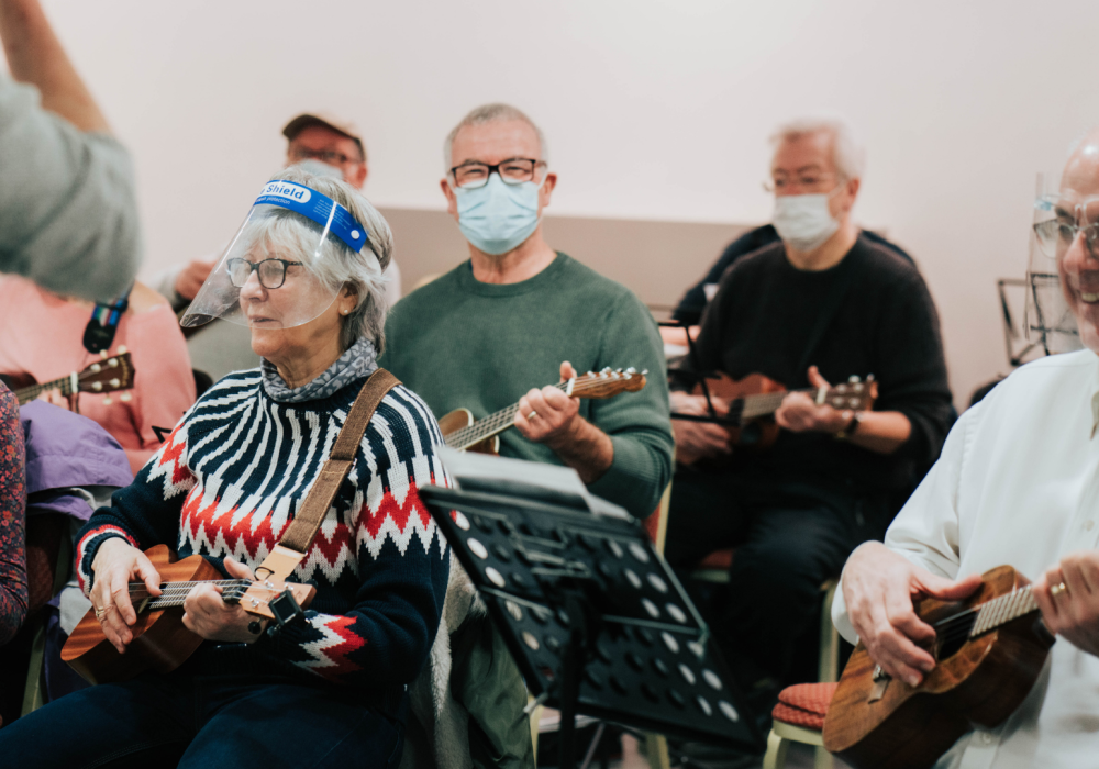 A group of people playing ukuleles, they are all smiling, and most are wearing face coverings.