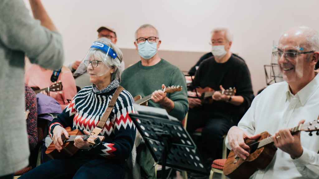A group of people playing ukuleles, they are all smiling, and most are wearing face coverings.