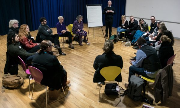 A group of 17 people sit in a circle (on chairs, some pink, some yellow) around a speaker standing next to a flip chart with notes on it. The room they're in - ARC's dance studio - has plain white walls, a wooden floor, and dark blue curtains down one side.