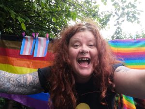 Julie Easley with her arms outstretched and a huge smile on her face. Behind her are various LGBTQ and trans flags hung over a line.