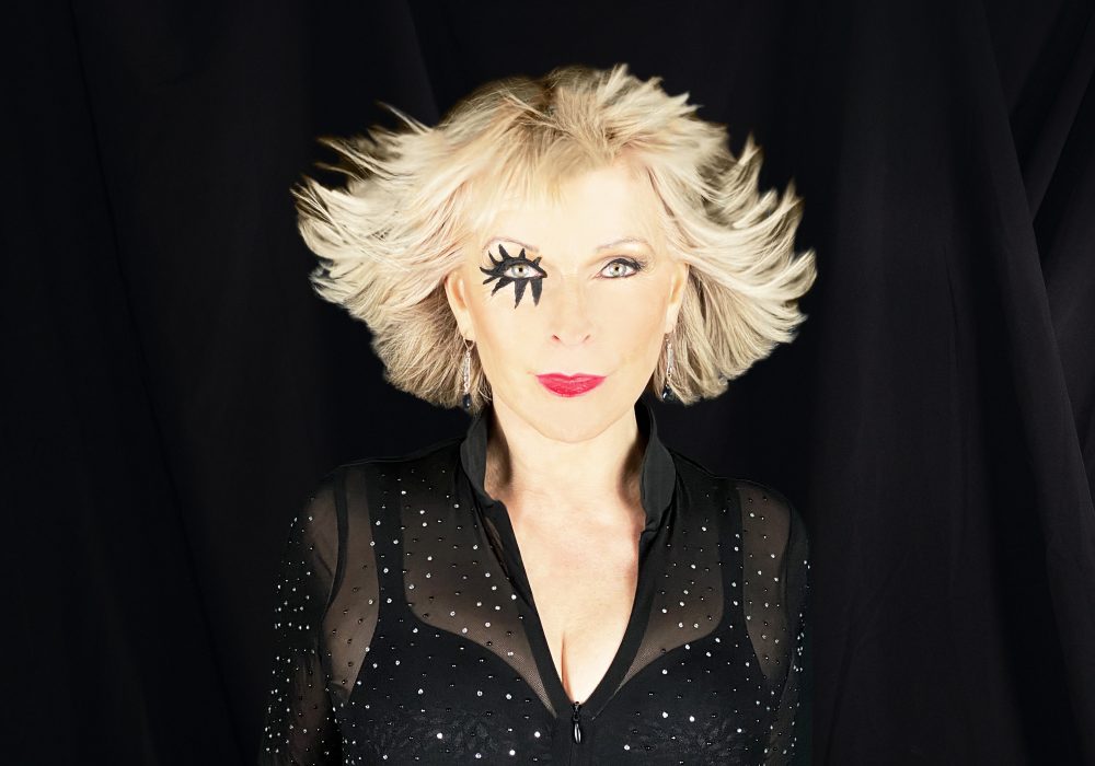 Singer Toyah stands against a black backdrop facing the camera. Her blonde hair is fanned away from her face, and she has drawn on black eyelashes on her left eye.