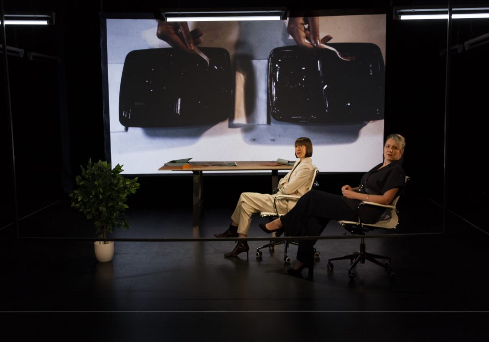Two female performers sit in office style chairs in front of a table on a black box style theatre set. They are both on the same side of the set and are in the same pose, looking side on towards the audience. There is a large pot plant on the front left of the set, and a projection screen in the background.