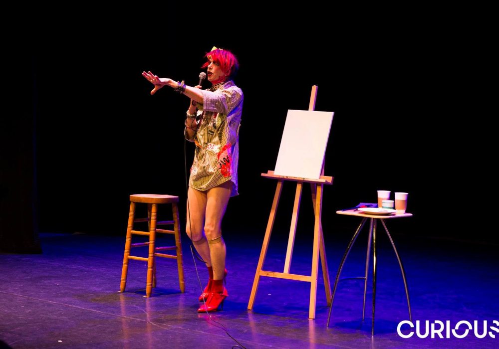 David Hoyle stands on stage wearing a long shirt, with bright red ankle boots. He has a tall stool to one side of him, and an easel and a small table to the other.