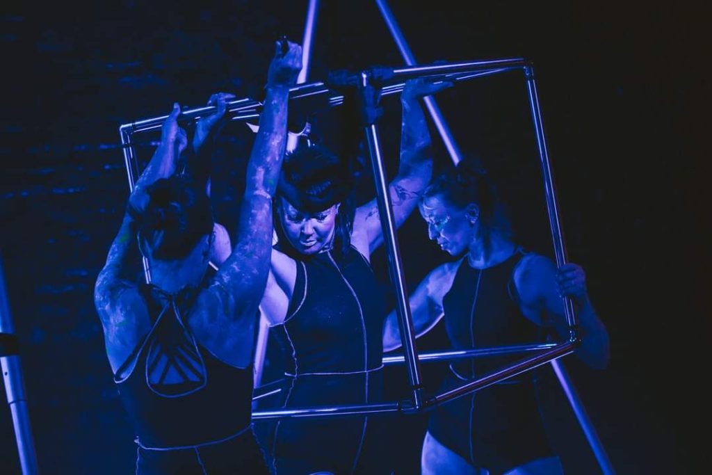 3 members of Uncaged Aerial Theatre in and around a square metal 'cage'