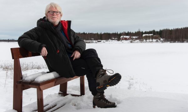 Artist Aidan Moseby (white hair, white beard, black framed rectangular glasses) sits on a small wooden snow-covered bench in a snowy field against a plain grey sly, He is wearing chunky walking boots and warm winter clothing (trousers and jacket).