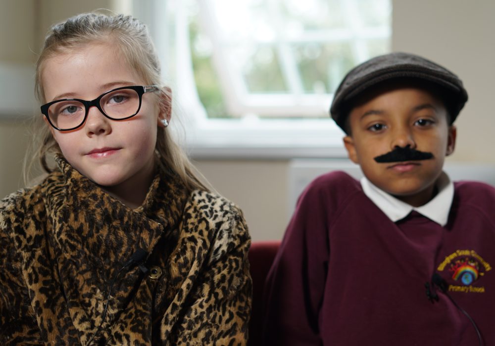 Two primary school children dressed up as themselves 50 years in the future. The boy is wearing a fake moustache.