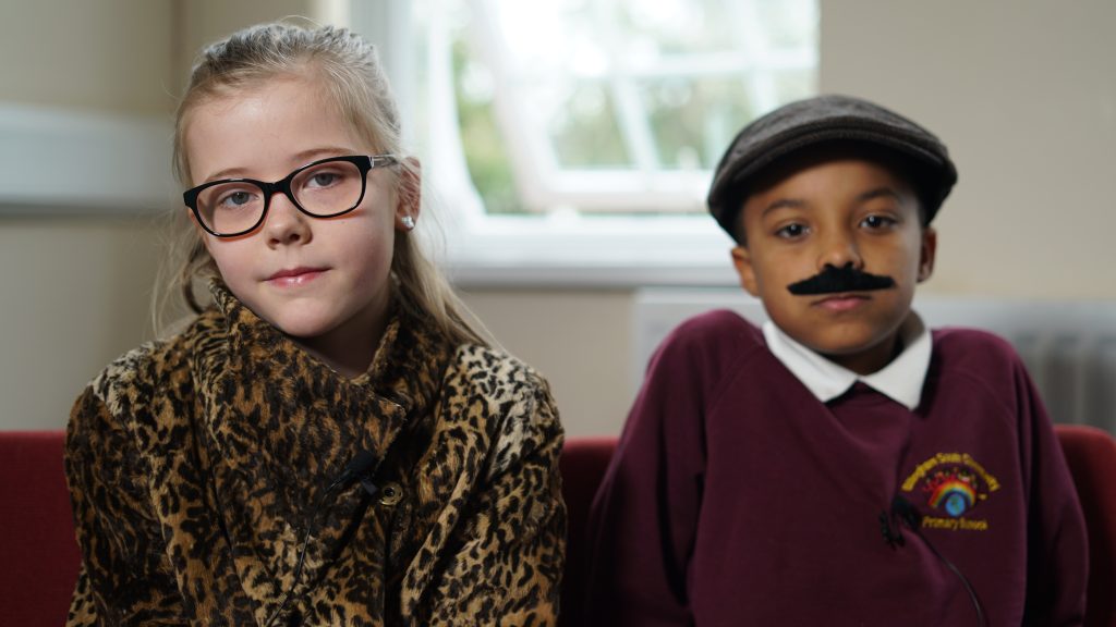 Two primary school children dressed up as themselves 50 years in the future. The boy is wearing a fake moustache.